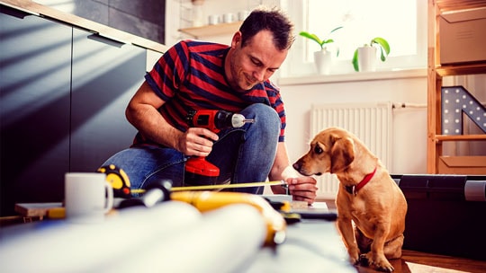 Man with drill in his hand measures objects on the floor while a dog watches.